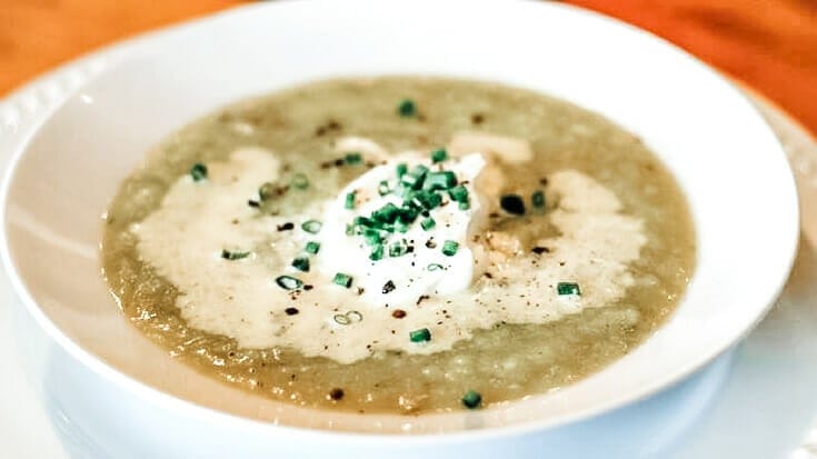 A closeup of the leek, garlic, and potato soup in a white shallow soup bowl with a dollop of sour cream and some chives sprinkled on top.