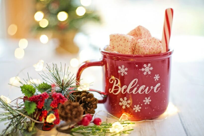 A red, christmas "believe" mug with peppermint coated marshmallows inside. Christmas greenery surrounds the mug and you can faintly see the Christmas tree in the background.