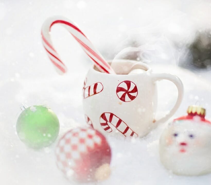 A white rounded mug with christmas candy pictures on it. Mug is filled with hot chocolate and a candy cane. Mug is surrounded with holiday ornaments.