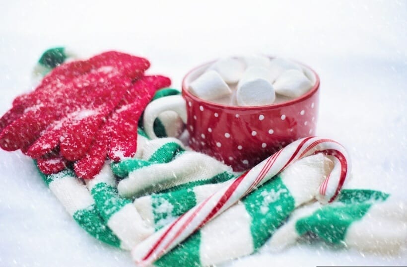 A red mug with white polka dots filled with cocoa and marshmallows. Mug is surrounded with red knit gloves, a green and white striped scarf, and a candy cane.