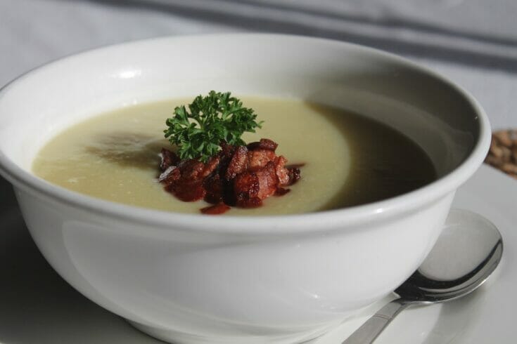 A deep white soup bowl filled with potato leek soup and garnished with bacon and a sprig of parsley.