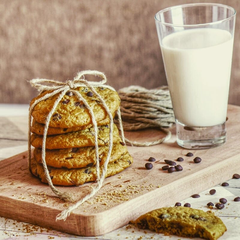 Cookies stacked up and tied with twine. A glass of milk in the background and chocolate chips spread around.