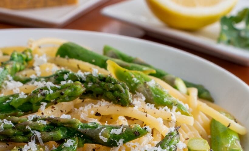 A close up of asparagus lemon pasta in a white bowl with a sliced meyer lemon in the background.