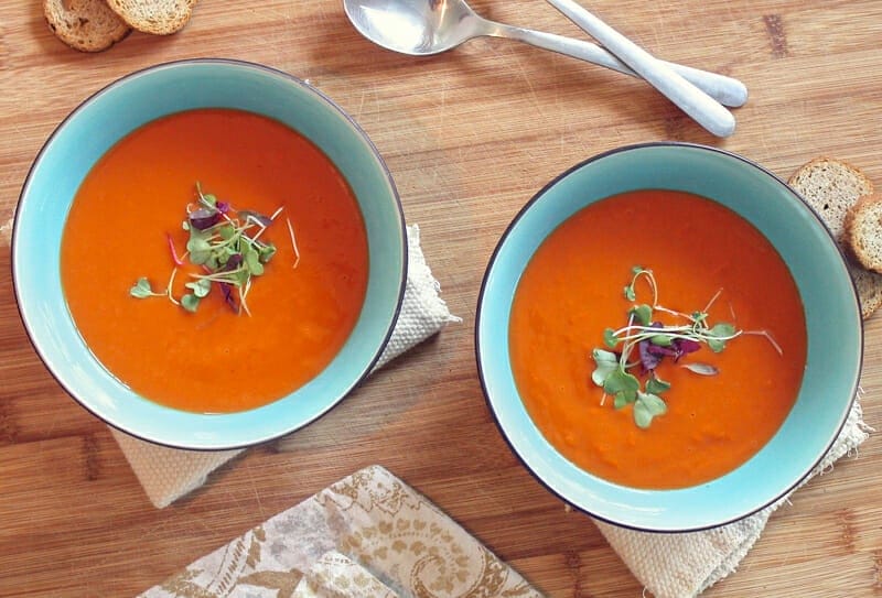 Two teal bowls of tomato soup garnished with spring greens on a wood table with burlap napkins.