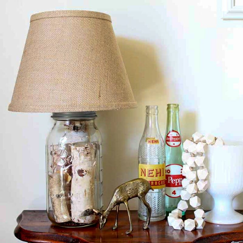 A large mason jar turned into a lamp with birch logs inside and a burlap shade.