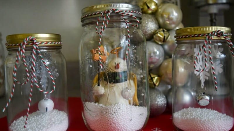 A christmas snowglobe with a snowman and snowflake ornaments inside mason jars and red and green ribbon tied around the lids.