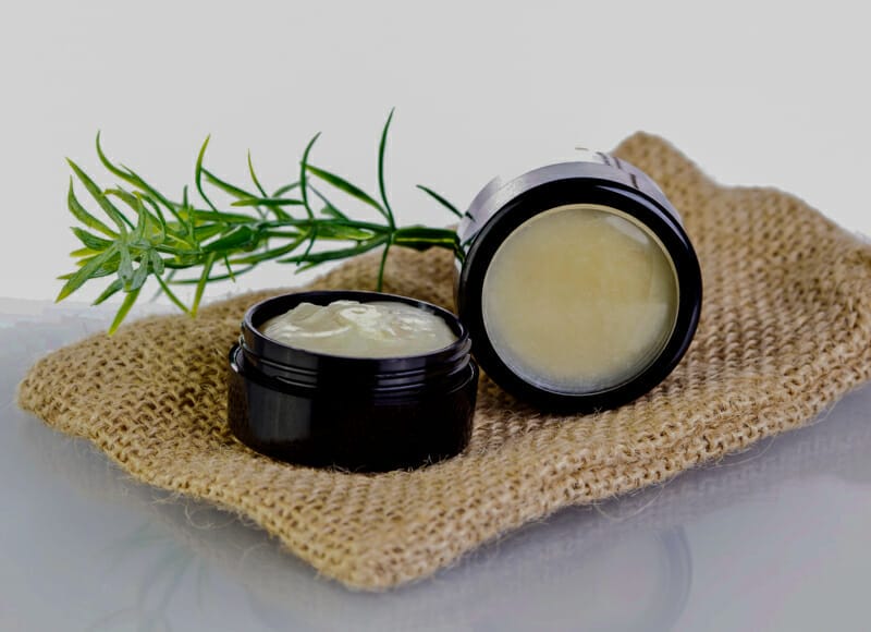 A plastic container of rosemary salve on burlap with a sprig of rosemary.