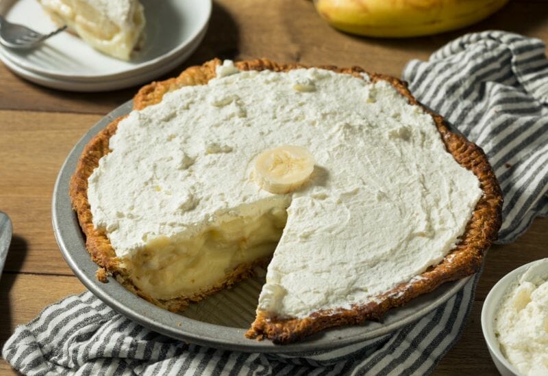 Banana cream pie with a homemade whipped cream topping on the counter with a slice cut from it.