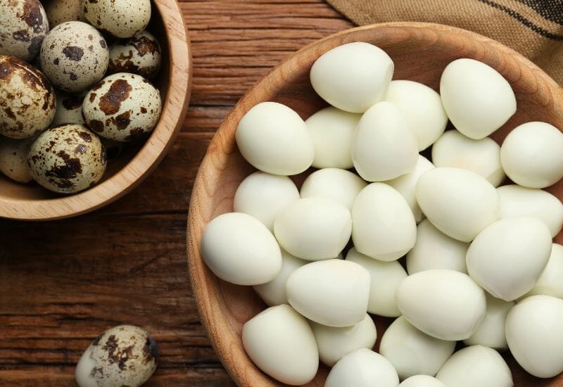 A wooden bowl of quail eggs and a second wooden bowl of peeled quail eggs.